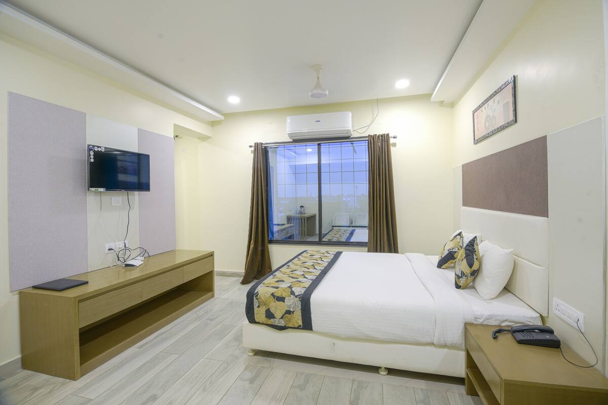 A hotel room with a bed, a TV mounted on the wall, a desk, and a window with curtains.
