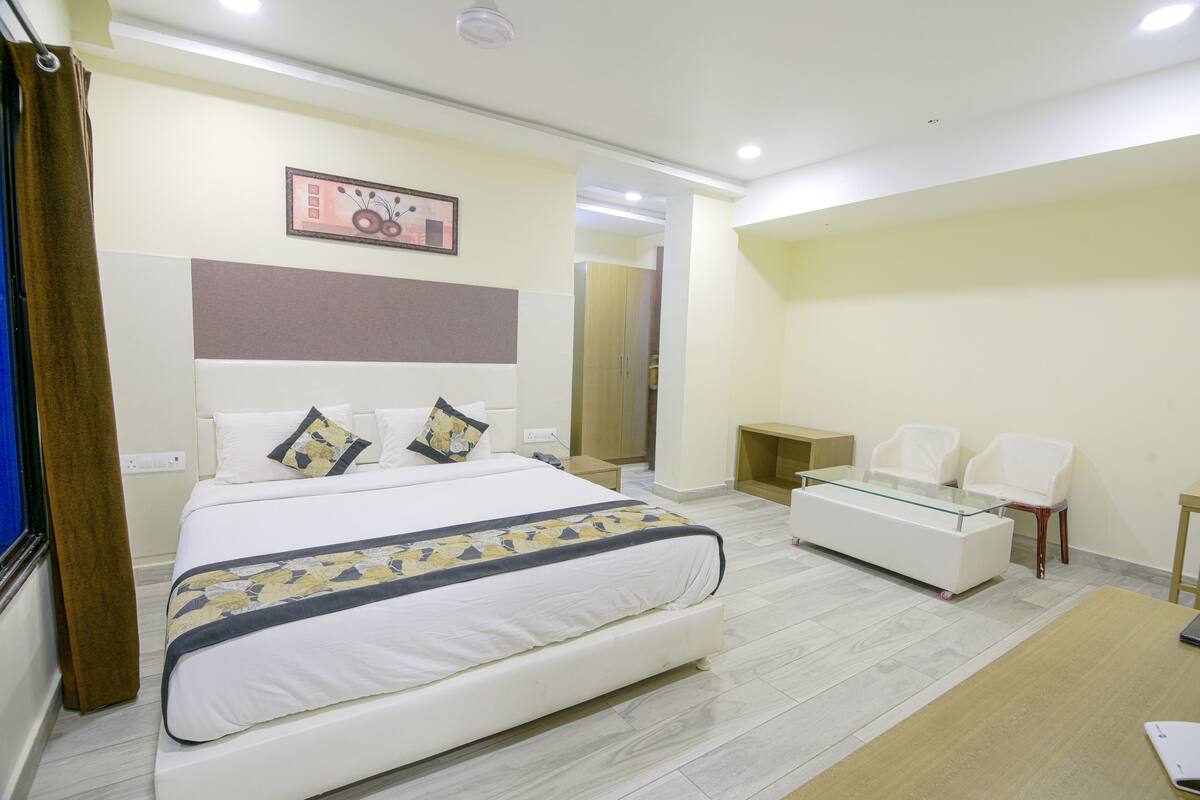 A hotel room with a large bed, a glass coffee table, two white chairs, a small table, and a mirror.