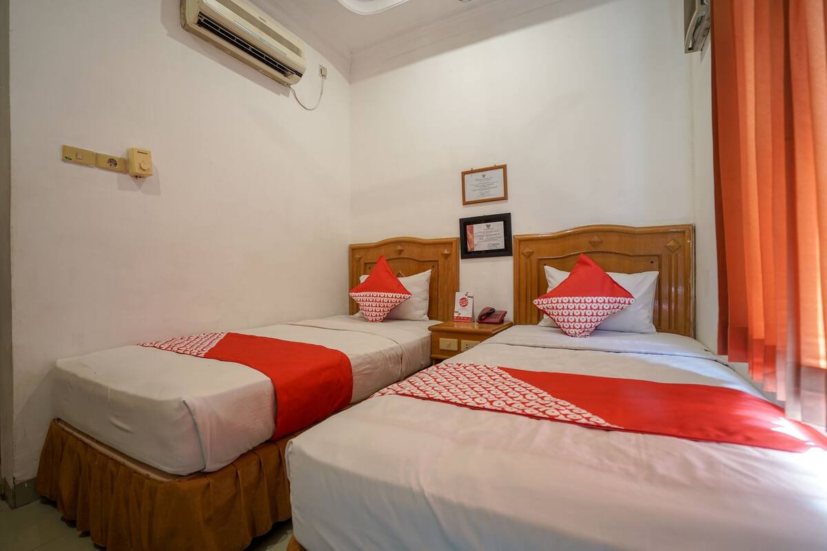 Habitación de hotel con dos camas, cada una con cabecero de madera, ropa de cama blanca y detalles en rojo. Hay una mesita de noche con un teléfono y un certificado enmarcado en la pared.