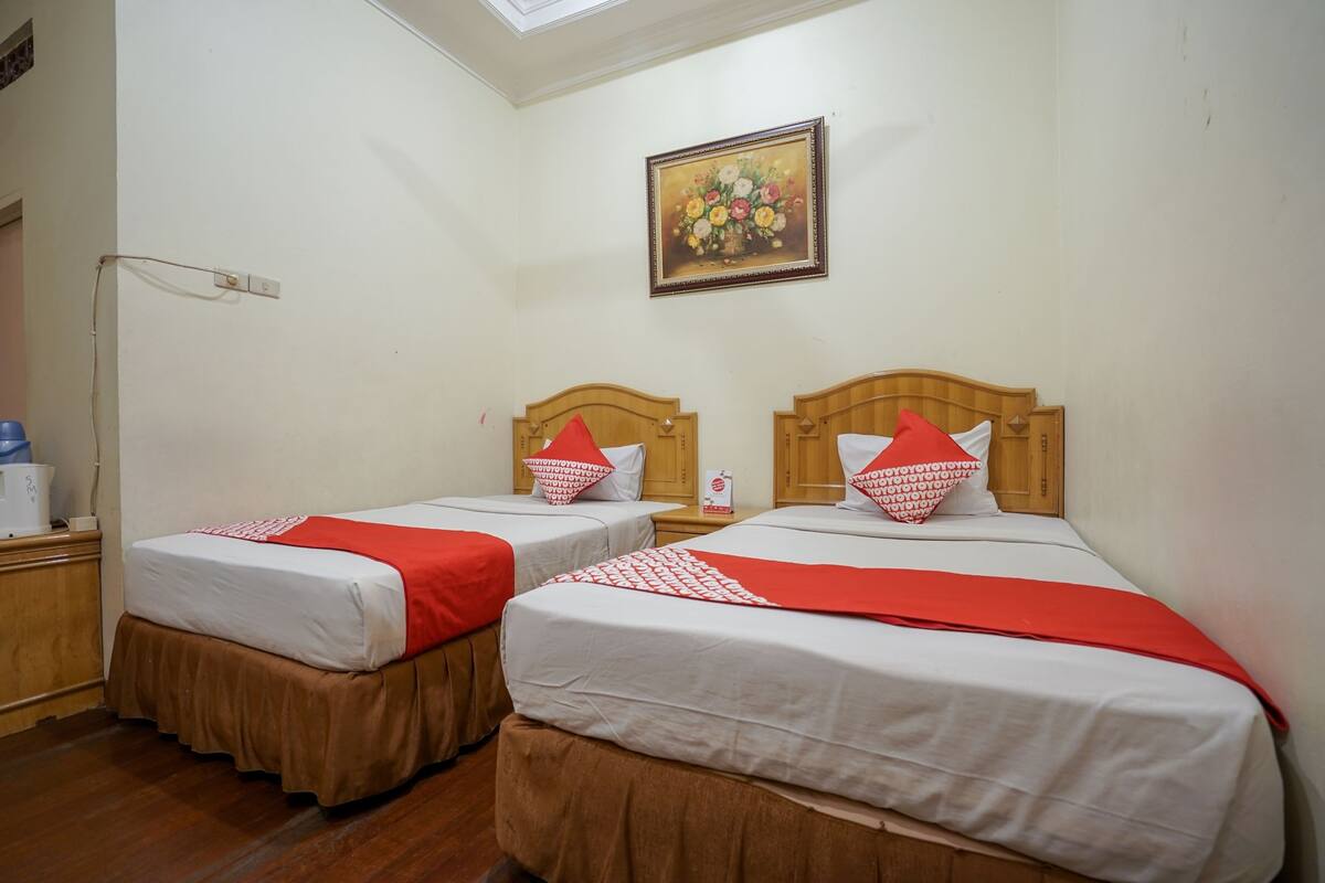 Habitación de hotel con dos camas, cada una con cabecero de madera, ropa de cama blanca y detalles en rojo. Un cuadro de flores cuelga de la pared sobre las camas.