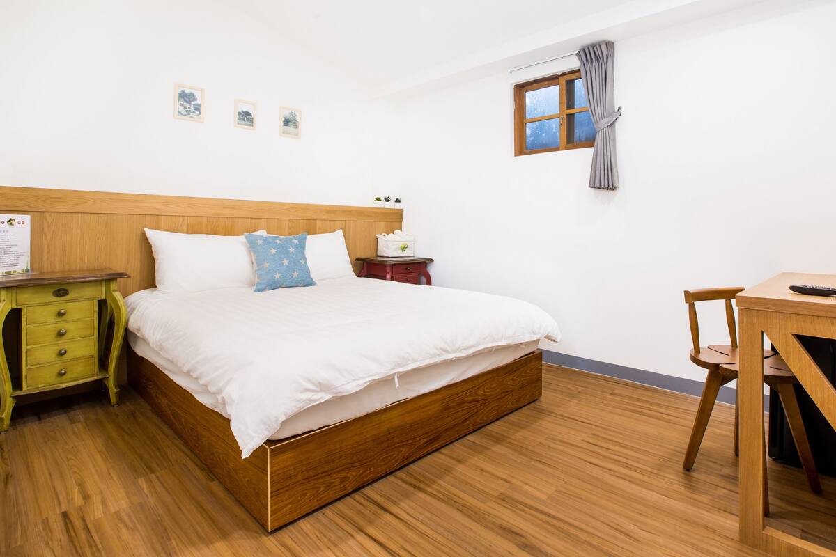 Une chambre à coucher bien rangée, avec un lit en bois, une table de chevet verte, un bureau en bois et une fenêtre avec des rideaux.