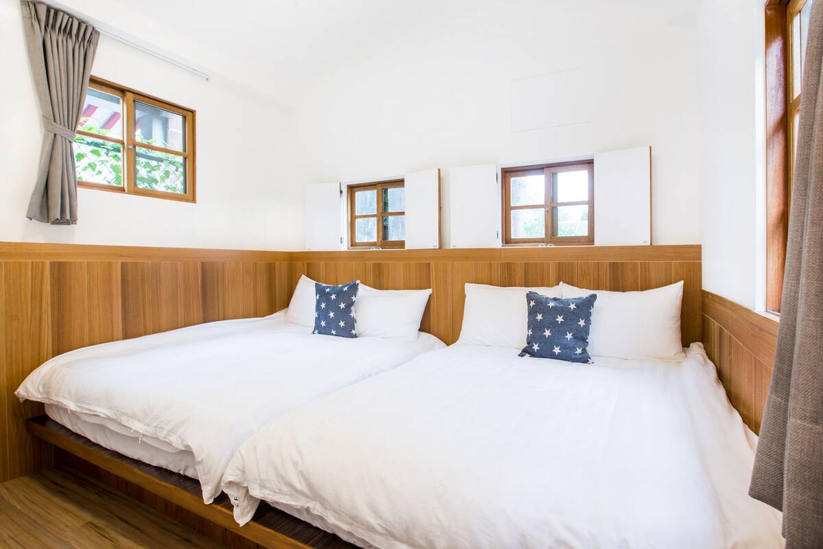 Une chambre d’hôtel avec deux lits, des têtes de lit en bois, du linge de lit blanc et une fenêtre avec des rideaux.