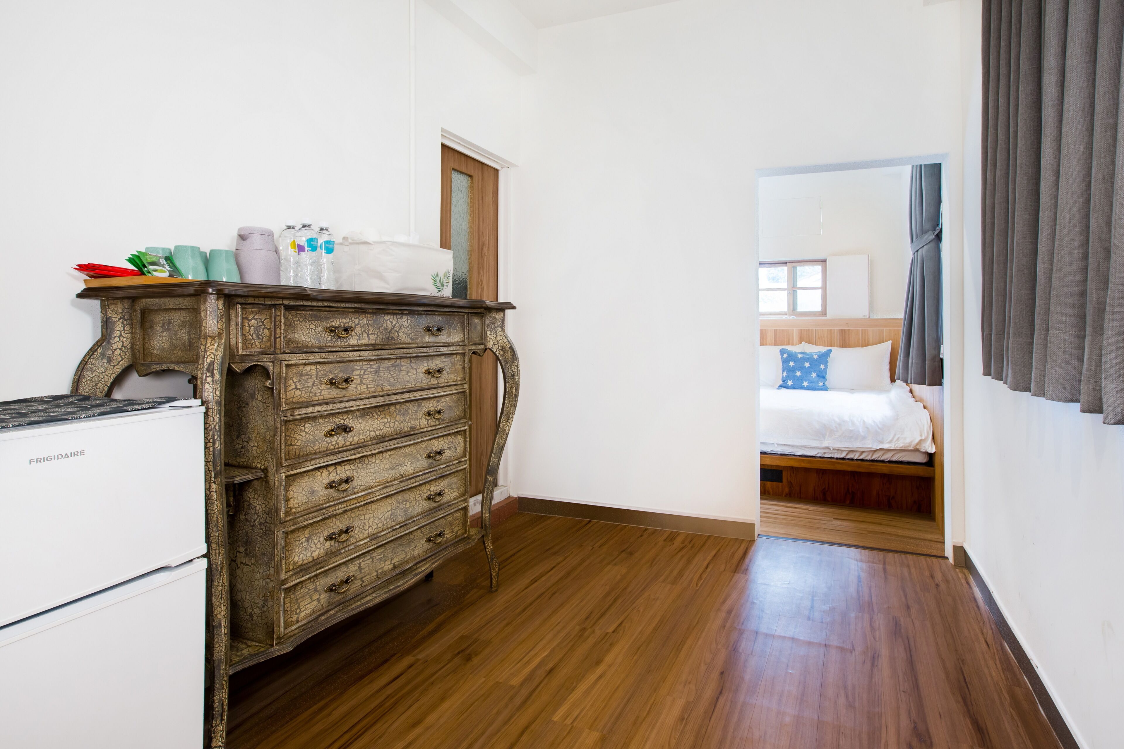 Une pièce avec une commode en bois, un lit avec un oreiller bleu et un réfrigérateur blanc.