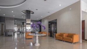Un hall d’entrée spacieux avec un sol en marbre, une table ronde centrale ornée d’un vase rempli de fleurs violettes, et des sièges orange disposés le long des murs.