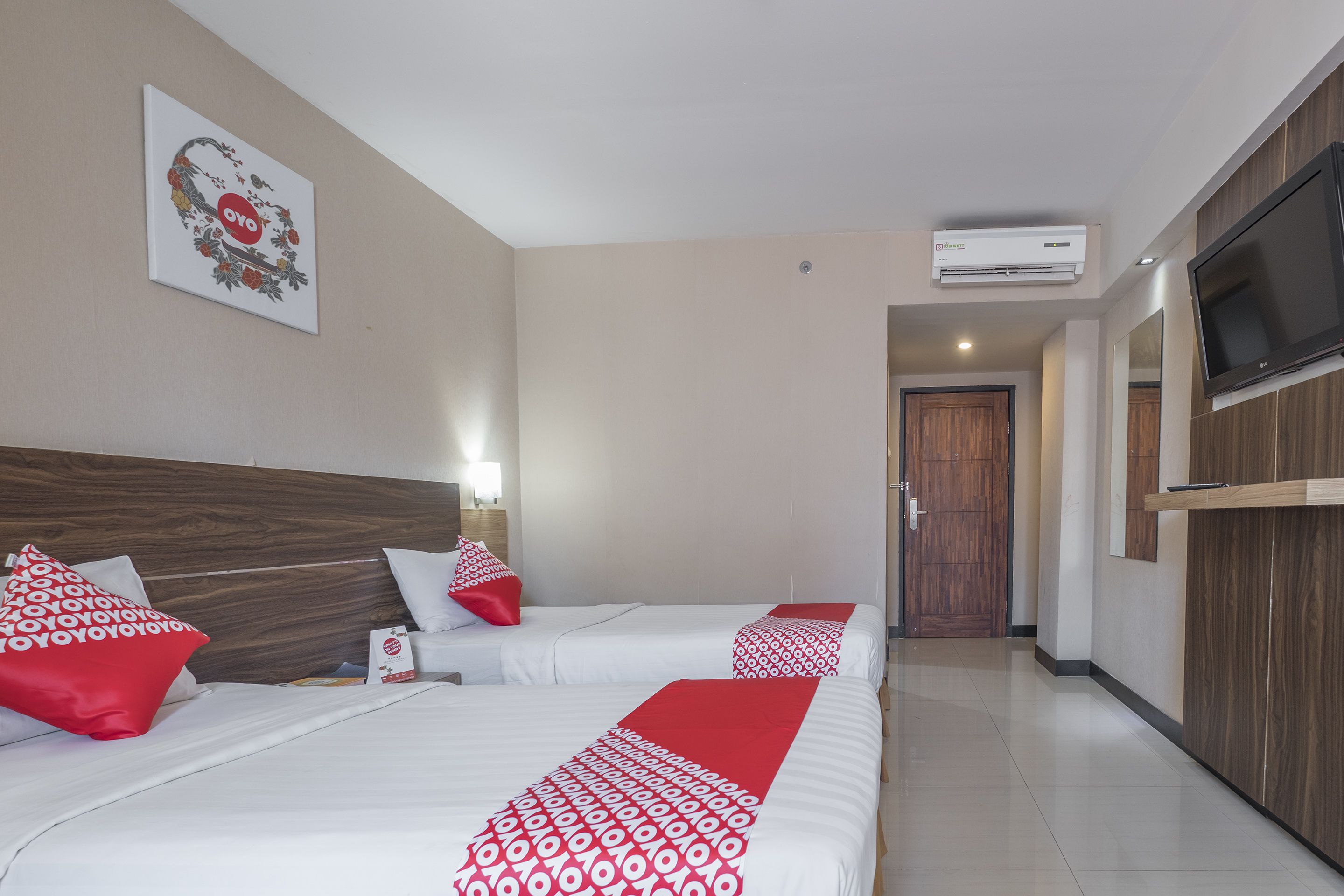 A hotel room with two beds, each with red and white patterned bedspreads, a wooden headboard, a wall-mounted TV, and an air conditioning unit.