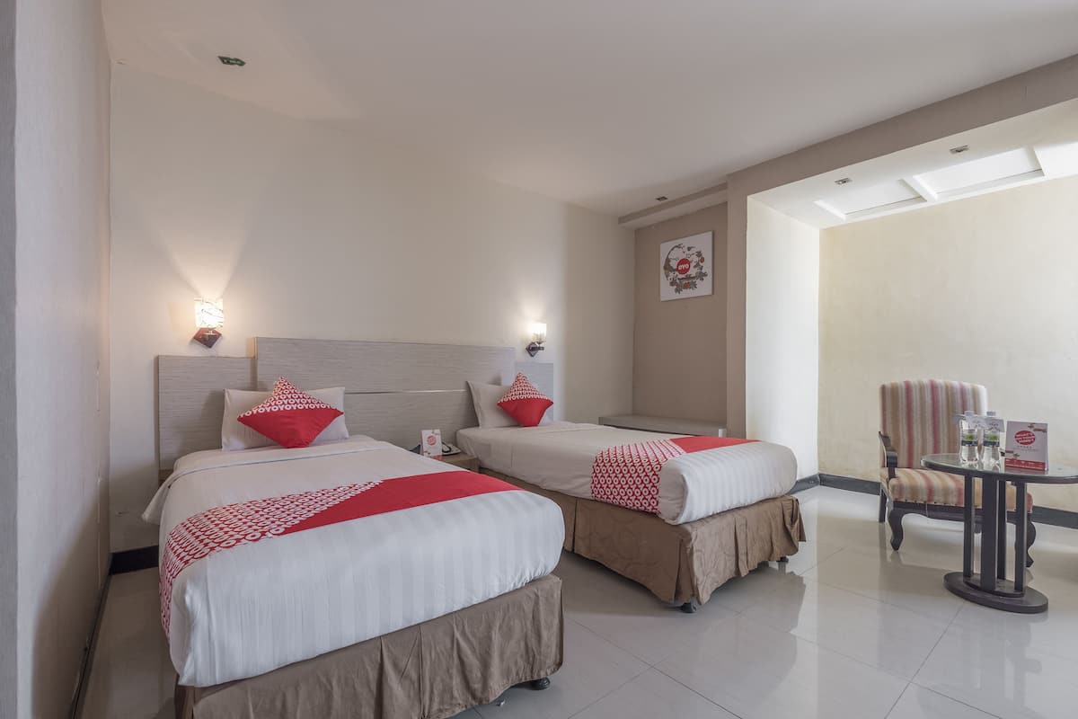 Une chambre d’hôtel avec deux lits, chacun recouvert d’une courtepointe rouge et blanche, doté d  ’une tête de lit en bois et agrémenté d’une table de chevet avec une lampe. On y trouve également une chaise et une petite table sur laquelle est posé un menu ou une brochure.