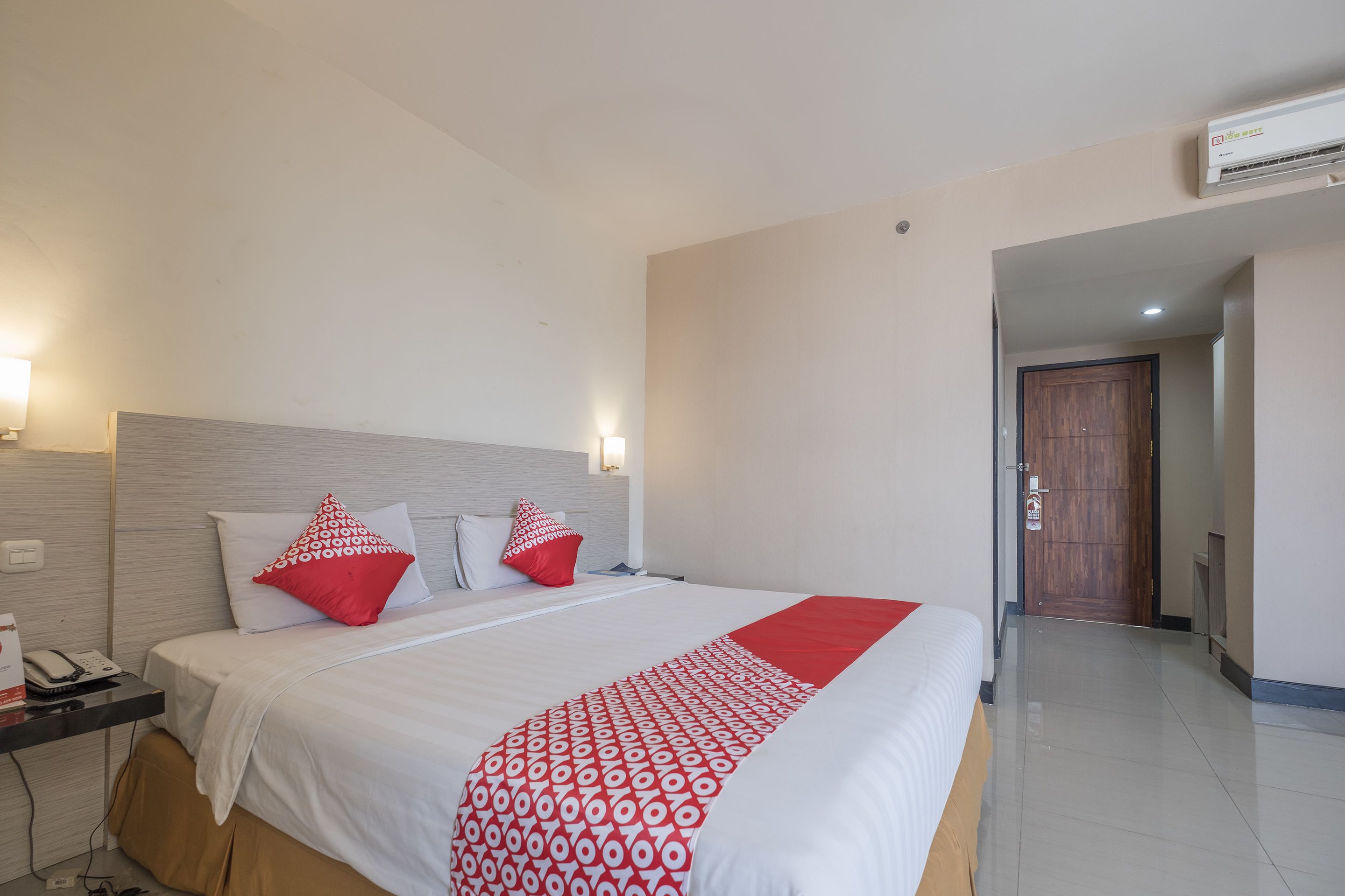 A hotel room with a large bed, white bedding, red and white patterned throw, and a wooden door in the background.
