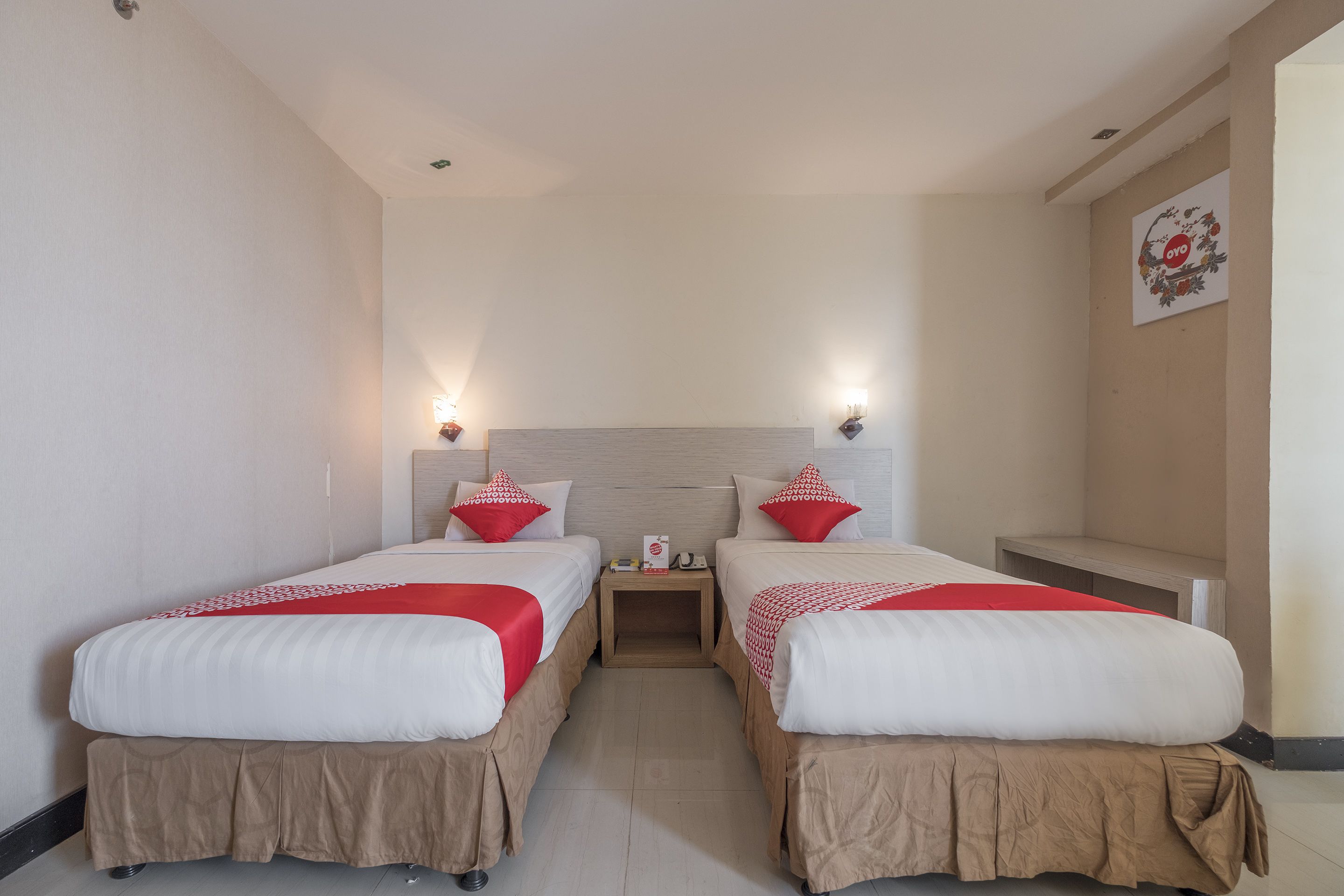 A hotel room with two beds, each with a red and white bedspread, a wooden nightstand, and a wall-mounted light fixture.