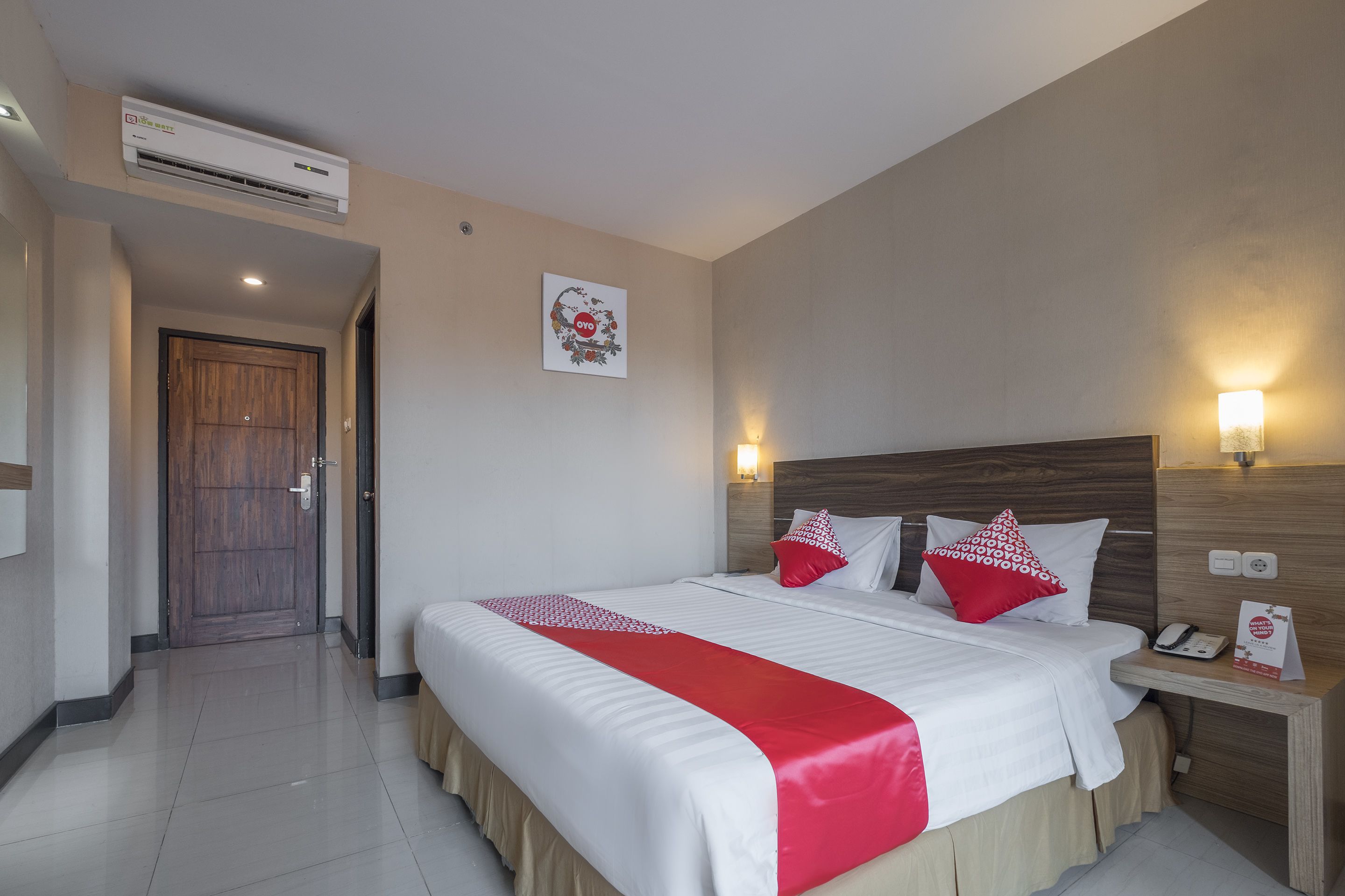 A hotel room with a large bed, a wooden headboard, a nightstand with a phone, and a wall-mounted air conditioner.