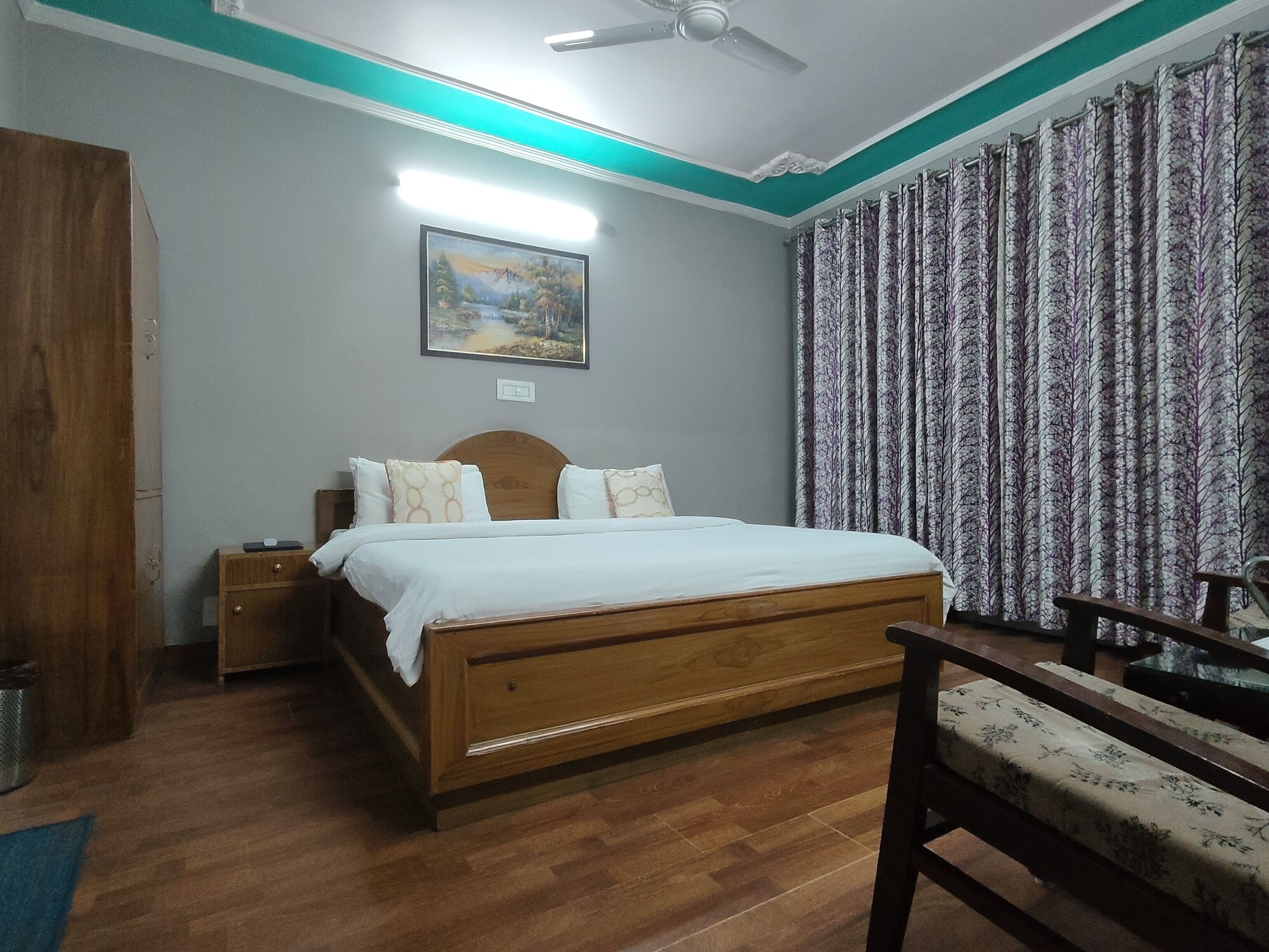 Un dormitorio con cama de madera, mesita de noche, una silla, un cuadro en la pared y cortinas.