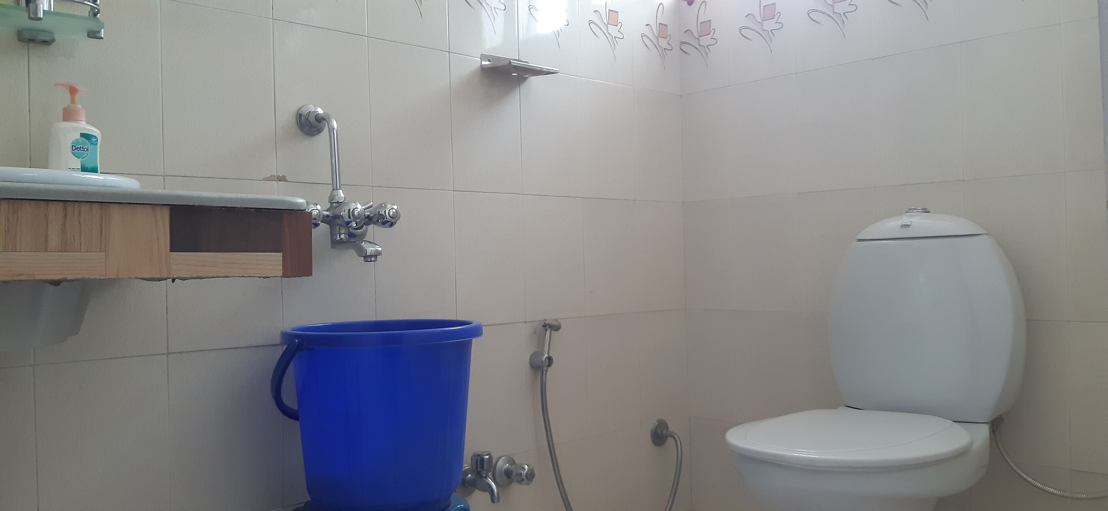 A bathroom with a toilet, a blue bucket, a showerhead, and a soap dispenser on a shelf.