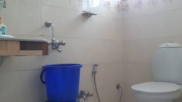 A bathroom with a toilet, a blue bucket, a showerhead, and a soap dispenser on a shelf.