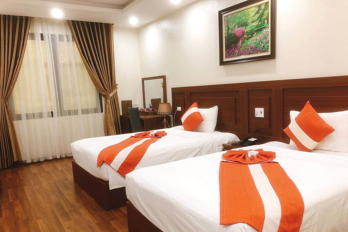 A hotel room with two beds, wooden headboards, a desk, a chair, a lamp, and a painting on the wall.