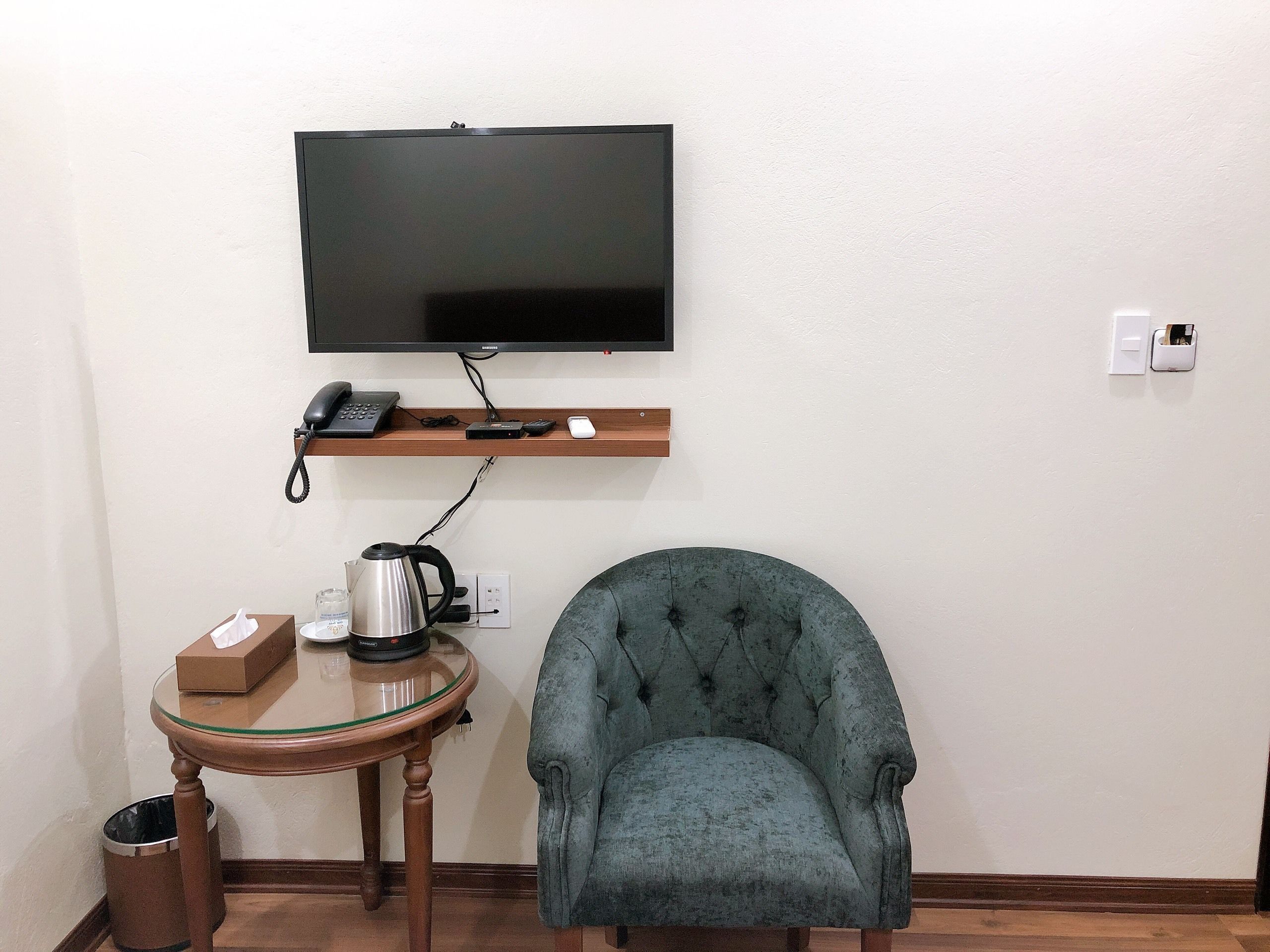 A room with a television mounted on the wall, a small wooden shelf with a telephone and a mouse, a round glass table with a kettle and a box, and a tufted armchair.
