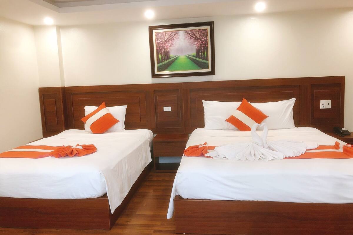 A hotel room with two single beds, wooden headboards, white bedding, orange accents, a framed picture on the wall, and a wooden floor.