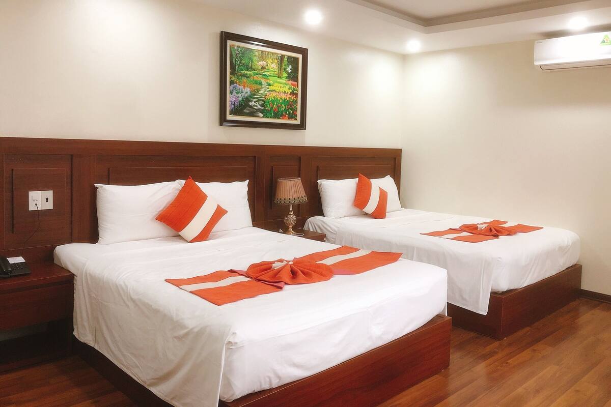 A hotel room with two beds, each with white bedding and orange accents, a nightstand with a lamp, and a painting of a garden on the wall.