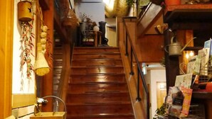 Una escalera de madera en un edificio con interior rústico. Hay estantes a la derecha con diversos objetos y una cesta a la izquierda.
