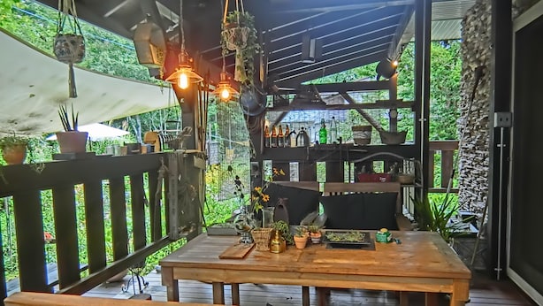 Una terraza de madera con una mesa y bancos, rodeada de plantas en macetas y luces colgantes.