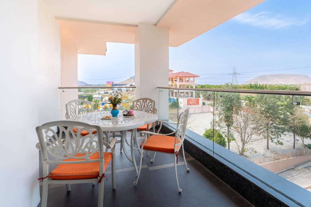 A balcony with a table and chairs, a vase with flowers, and a view of buildings and trees.