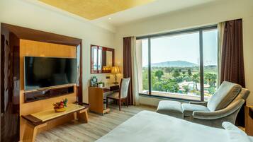 A hotel room with a large flat-screen TV, a desk with a lamp, a comfortable chair, and a bed. There is a large window offering a view of the outdoors.