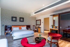 A modern hotel room with a large bed, a desk, a red chair, and a dining area with a table and chairs.
