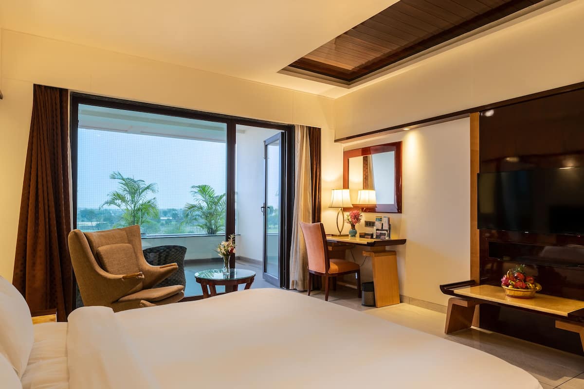 A hotel room with a bed, a desk, a chair, a TV, and a balcony with a view of palm trees.