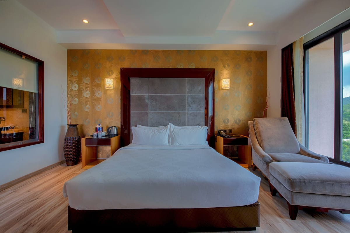 A hotel room with a large bed, a chair, a nightstand, a vase, and a window with curtains.