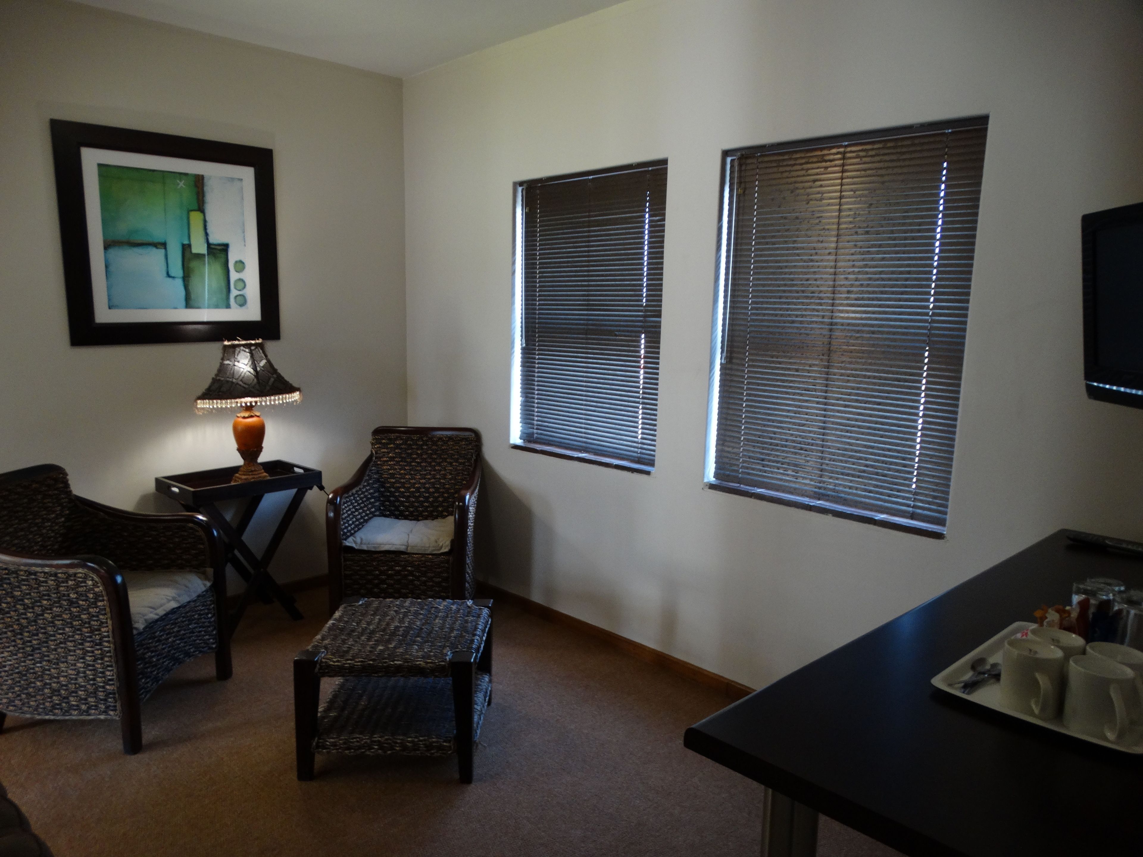 Una sala de estar con dos sillones, una mesita con una lámpara y una mesa de centro. Hay dos ventanas con persianas, un cuadro enmarcado en la pared y un televisor de pantalla plana sobre un mueble.