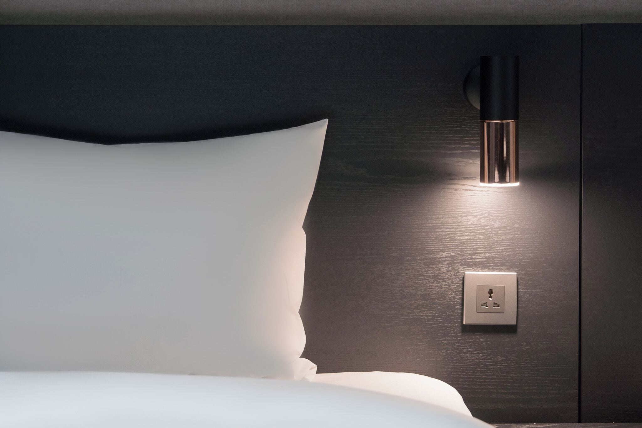 A modern bedroom with a bed, a wall-mounted light fixture, and an electrical outlet.
