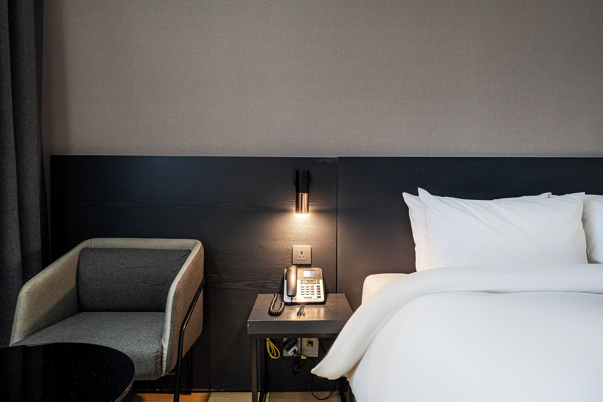 Une chambre d’hôtel comprenant un lit, une table de chevet avec un téléphone, une chaise et une lampe fixée au mur.
