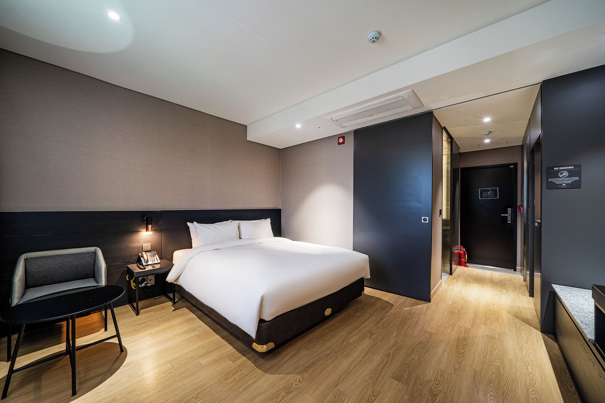 Una habitación de hotel moderna con una cama grande, mesita de noche, lámpara y una pequeña silla.