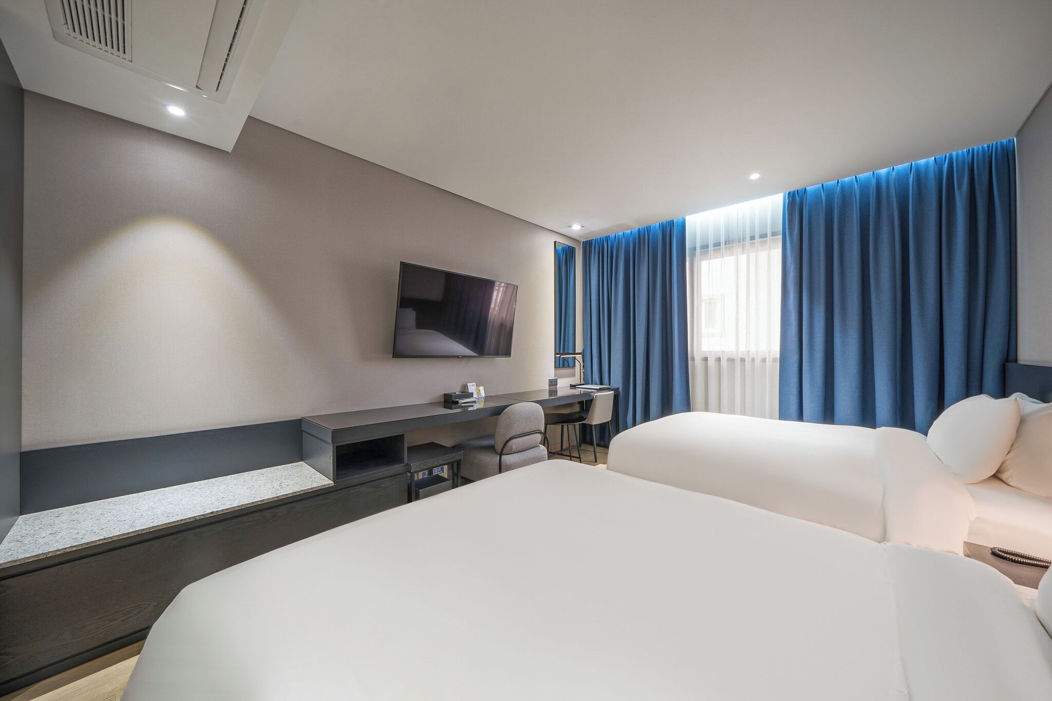 A modern hotel room with a large bed, a desk with a chair, a flat-screen TV mounted on the wall, and a window with blue curtains.