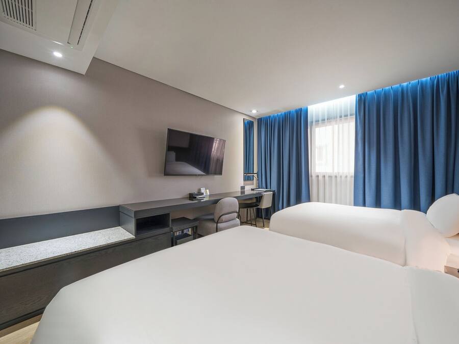 Une chambre d’hôtel moderne dotée d’un grand lit, d’un bureau avec une chaise, d’un téléviseur à écran plat fixé au mur et d’une fenêtre avec des rideaux bleus.