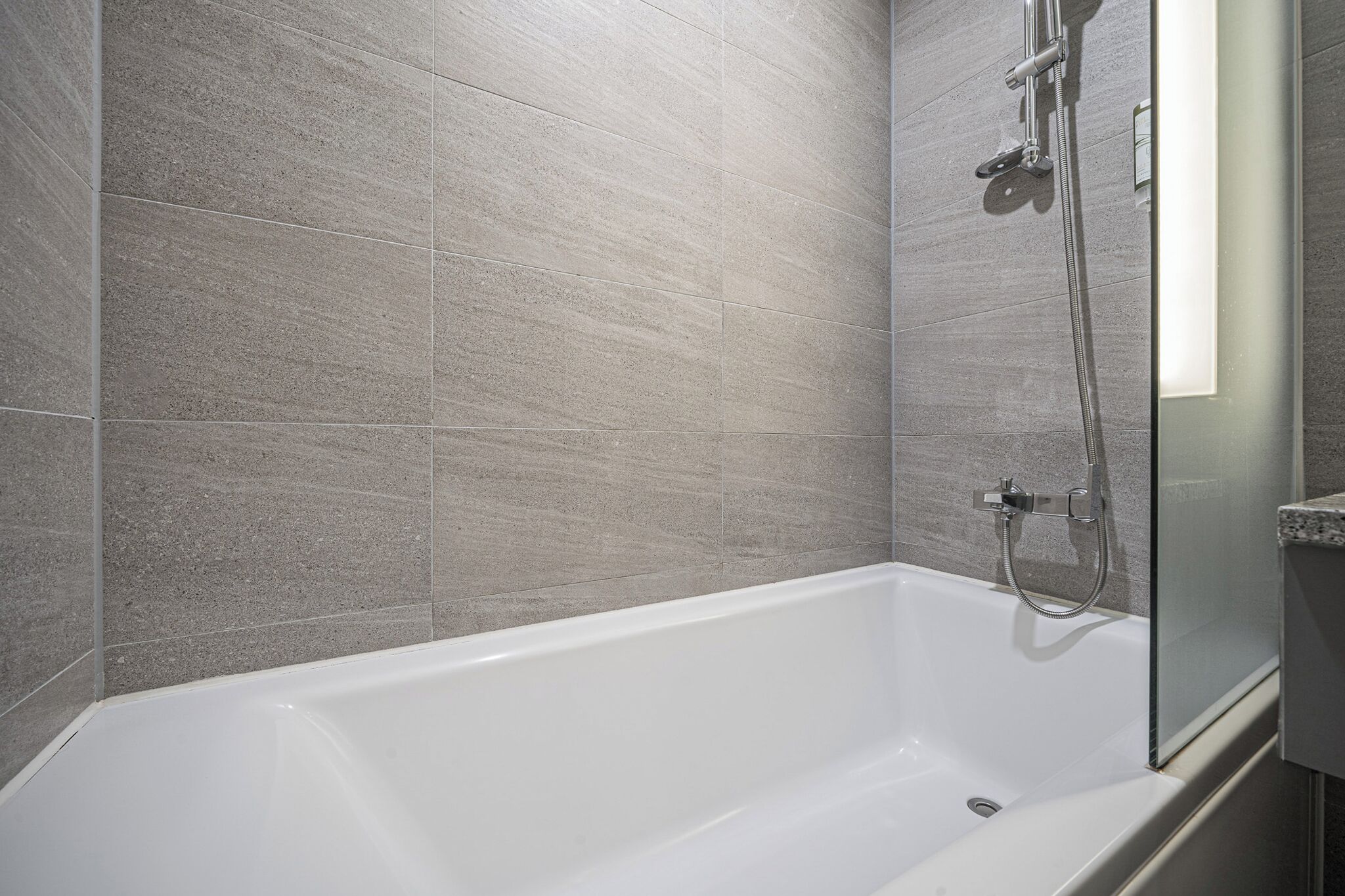 A modern bathroom with a white bathtub, a glass shower enclosure, and grey tiled walls.