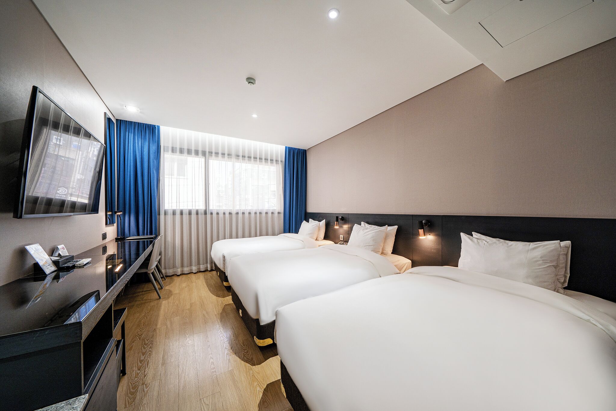 A hotel room with two beds, a desk with a flat-screen TV, and a window with blue curtains.