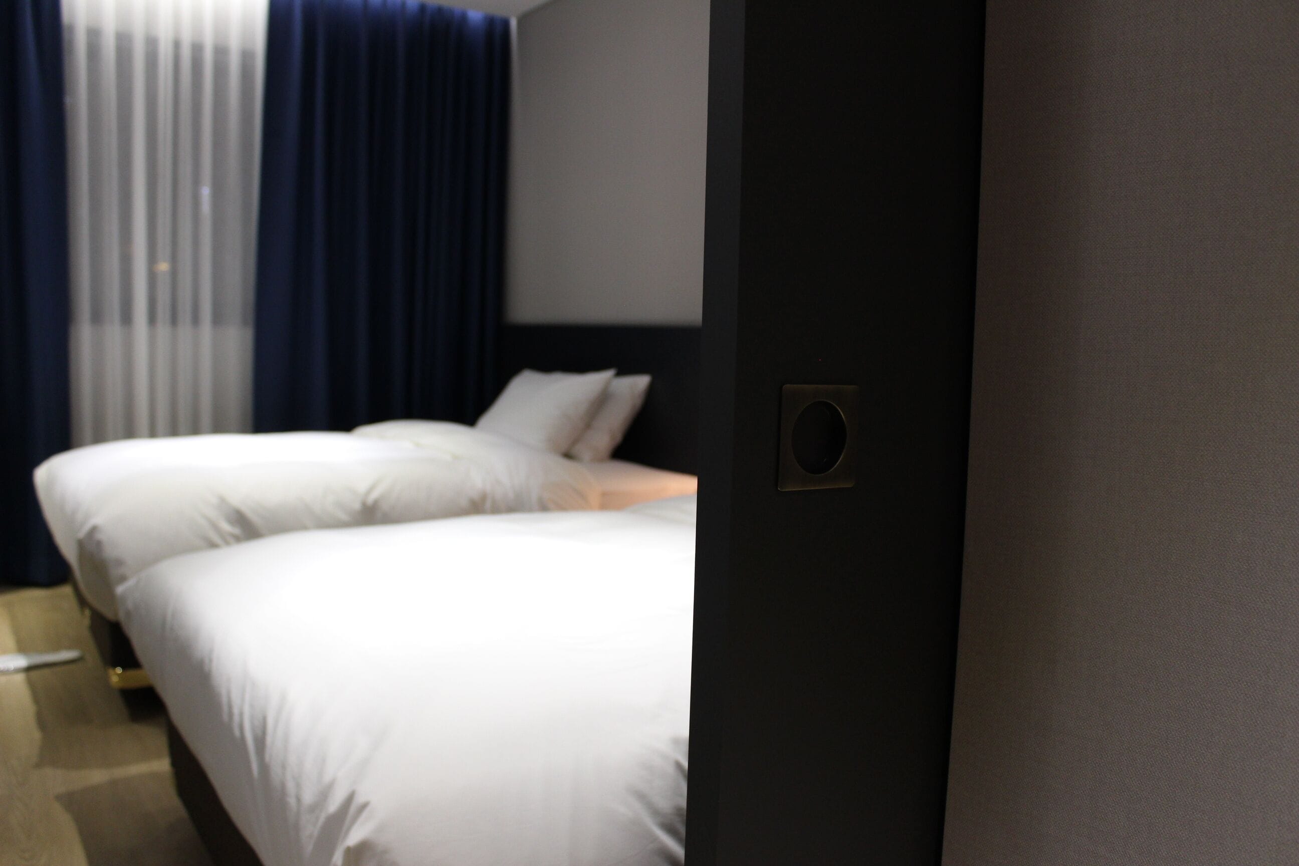 A hotel room with two beds, a dark-colored wall, and a window with blue curtains.