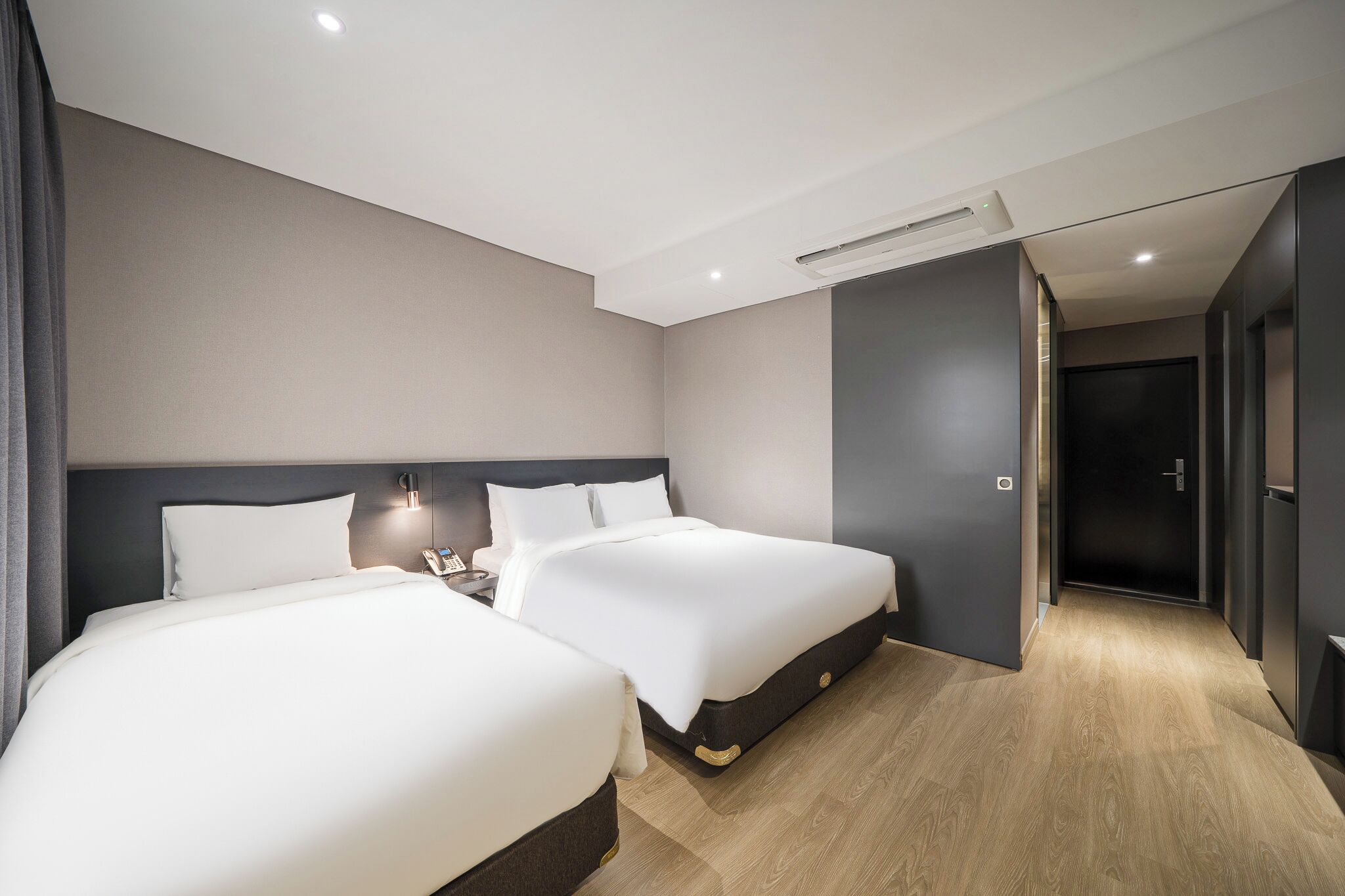 Habitación de hotel con dos camas, una mesita de noche con lámpara, un aire acondicionado instalado en la pared y una puerta que da a otra habitación.