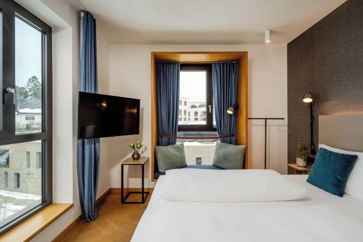 A hotel room with a large bed, a TV, a window with curtains, and a view of buildings outside.