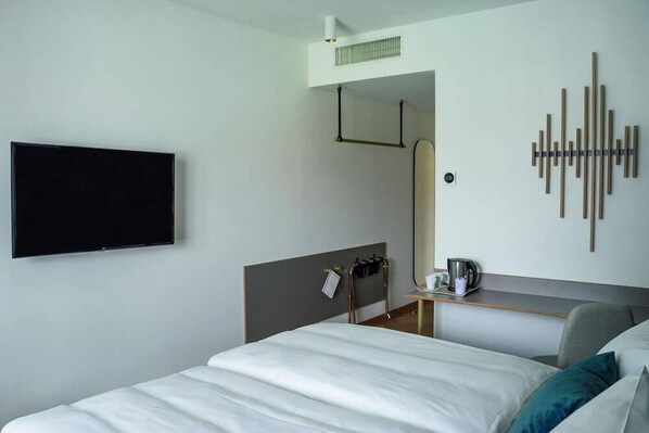 Una habitación de hotel con una cama, un televisor de pantalla plana montado en la pared, una cafetera sobre una mesita y un adorno en la pared.