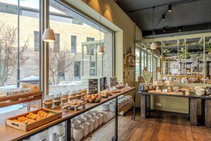 A café with a large window, wooden tables, and various food items displayed. - Welcome Hotel Kronberg Musikquartier (Kronberg)
