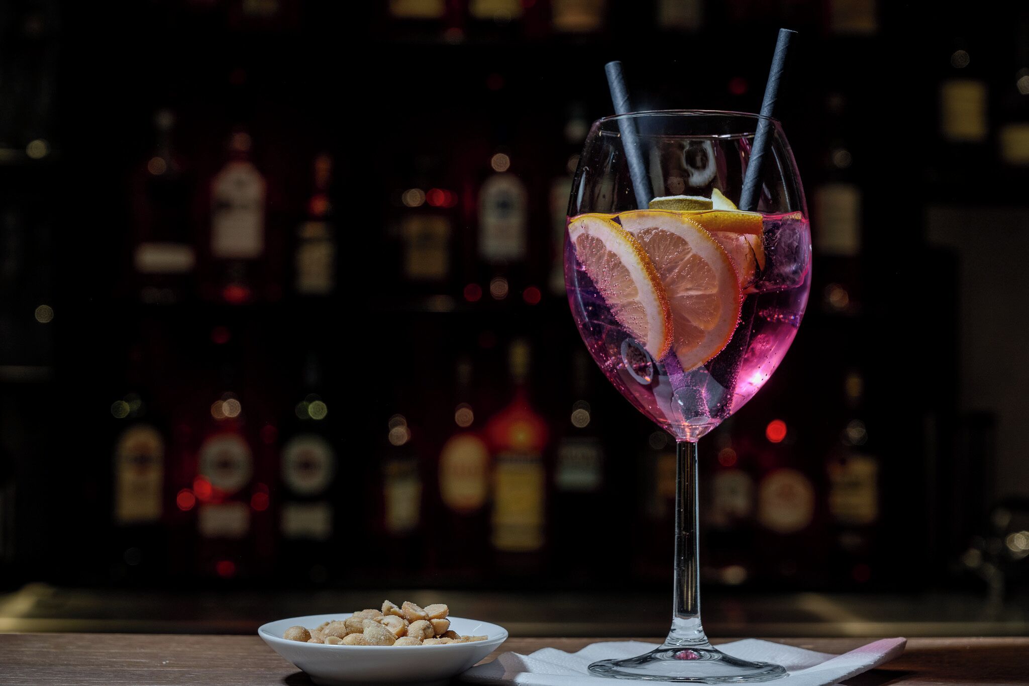 A cocktail glass with a straw, garnished with citrus slices, on a bar counter with a blurred background of bottles.
