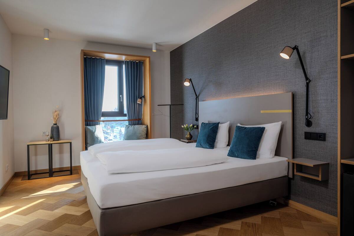 Une chambre d’hôtel moderne avec un grand lit, des lampes de chevet, une petite table avec un vase et une vue par la fenêtre, qui est dotée de rideaux.