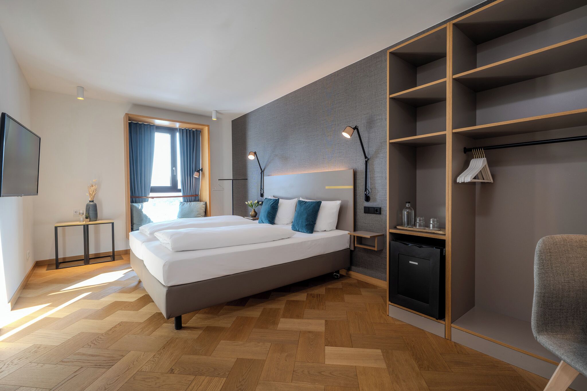 A modern hotel room with a large bed, bedside lamps, a wardrobe, and a desk with a chair.