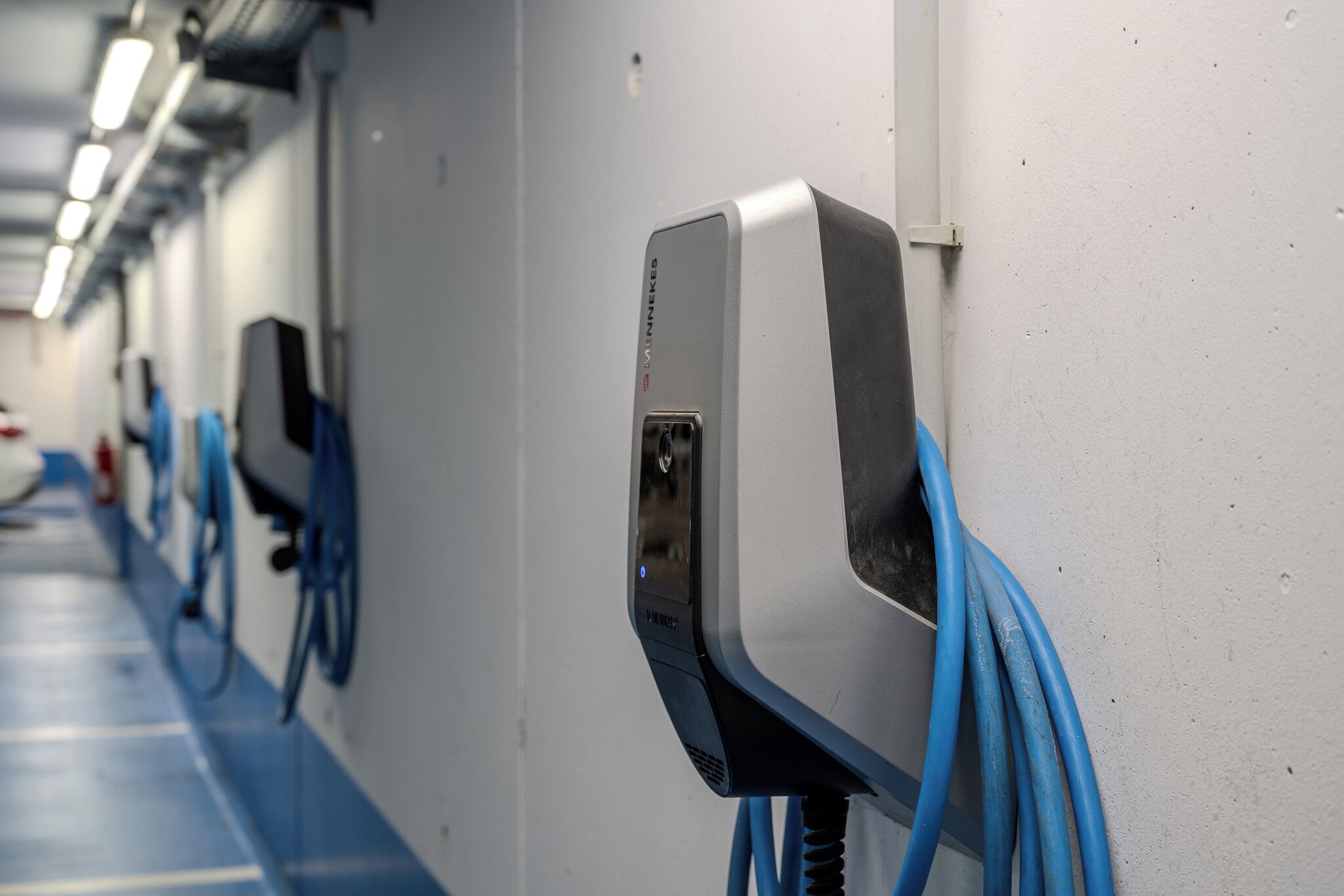 A row of charging stations with blue cables, a white wall, and a blue floor.