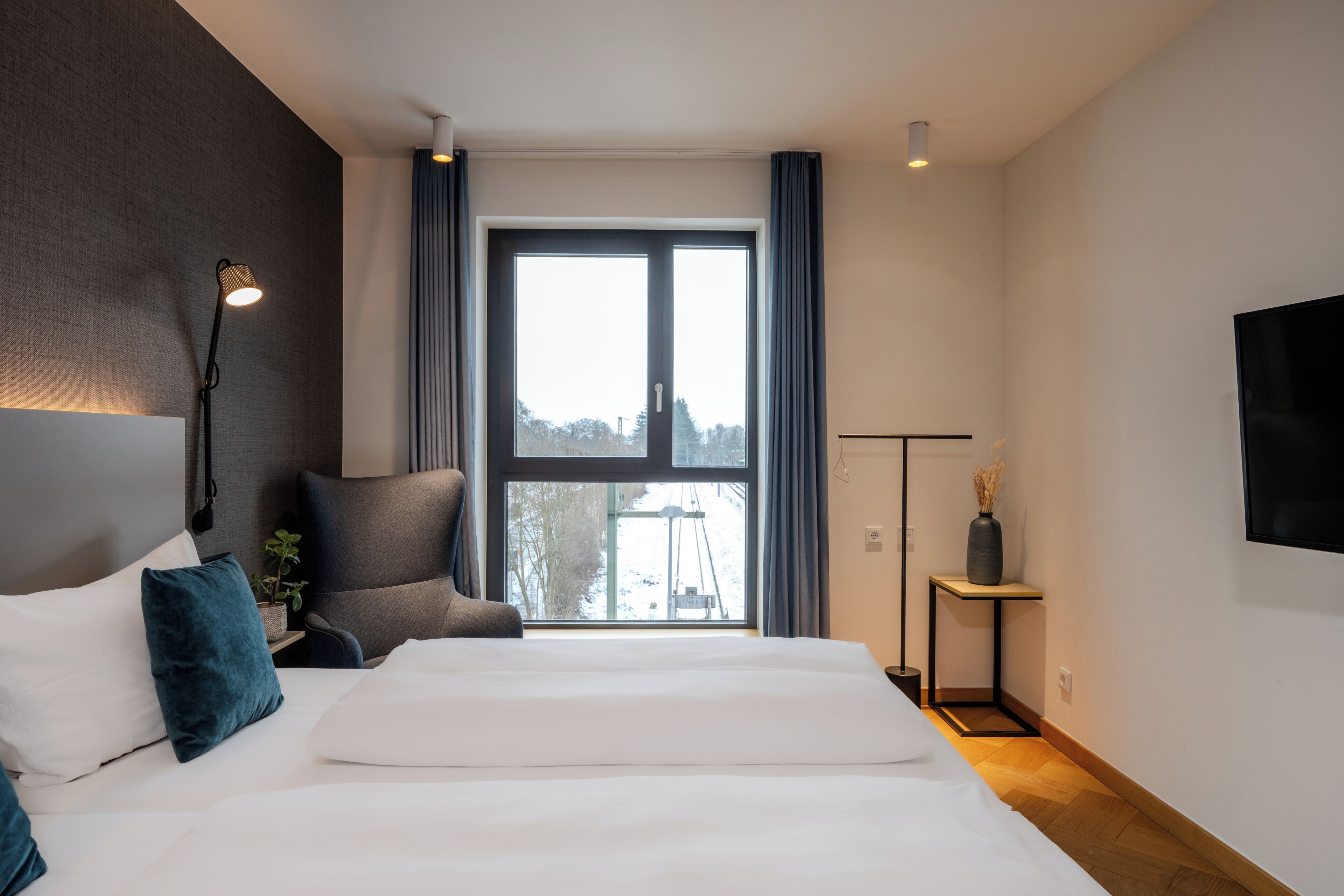 A modern bedroom with a large bed, a chair, a bedside table, a lamp, and a view of the outdoors.
