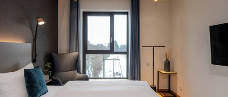 A modern bedroom with a large bed, a chair, a bedside table, a lamp, and a view of the outdoors.