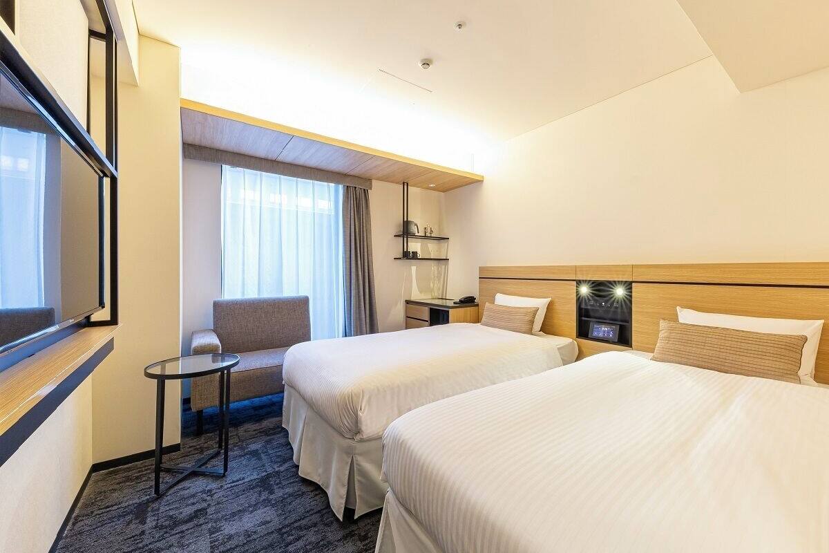 A hotel room with two beds, a small table, a chair, a TV, and a window with curtains.