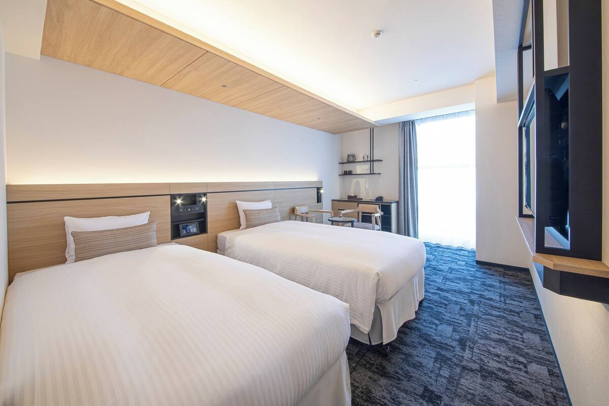 A hotel room with two beds, a desk, a chair, a TV, and a large window.