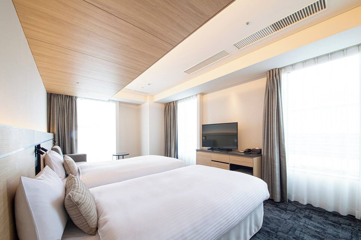 A hotel room with two beds, a desk with a TV, and large windows with curtains.