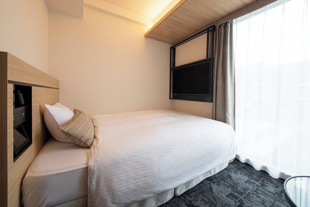 A hotel room with a bed, a TV mounted on the wall, a small table, and a window with curtains.